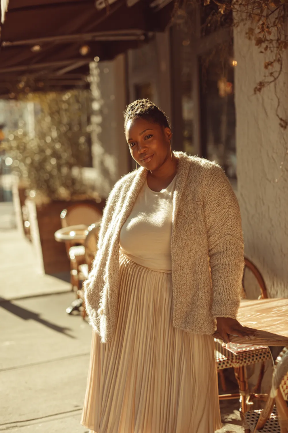 Plus size woman in a pleated midi skirt and knit cardigan standing outside a café in warm sunlight