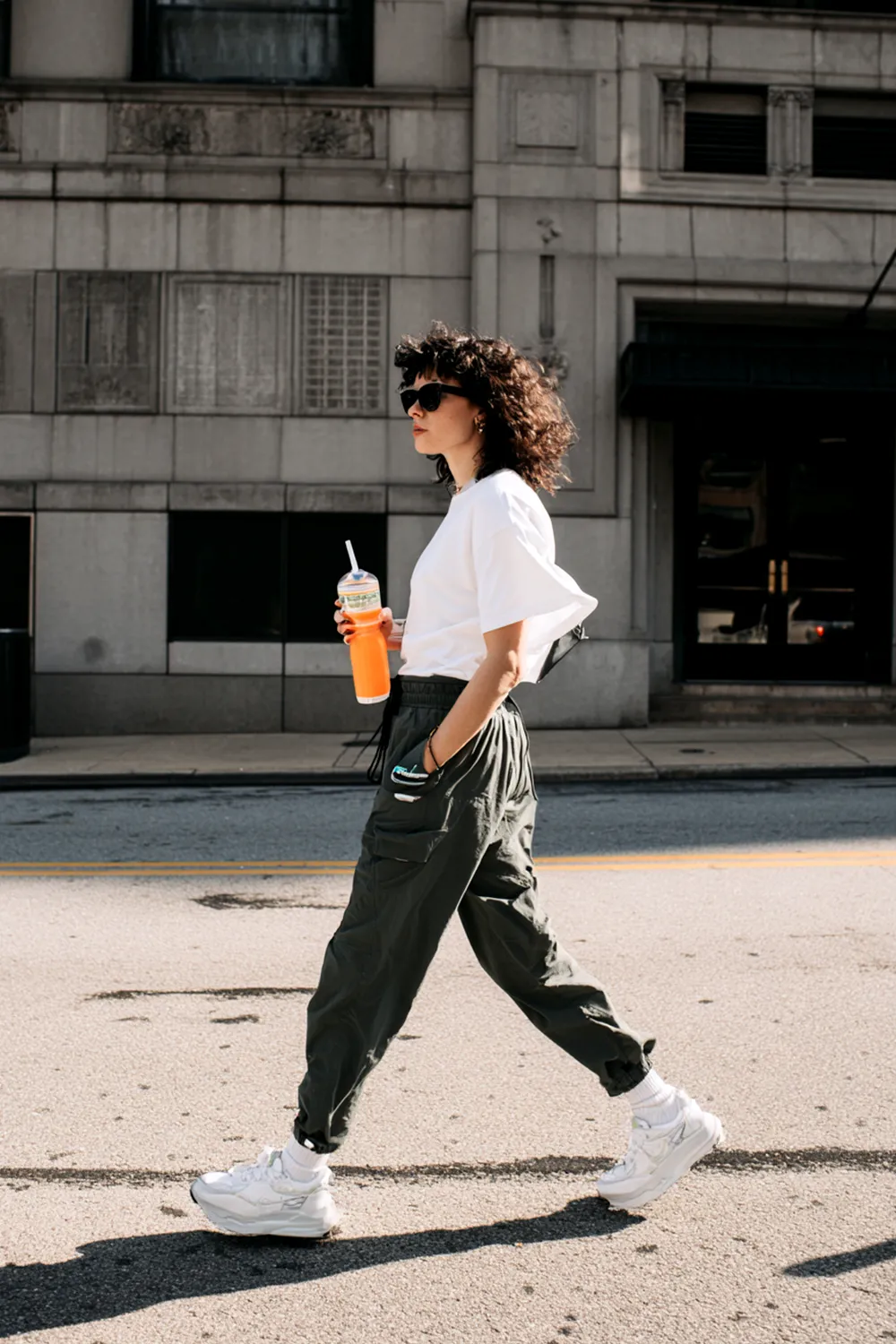Candid side-profile photo of a woman walking in white chunky sneakers, dark cargo pants and a white tee, holding a bright orange drink on a city street
