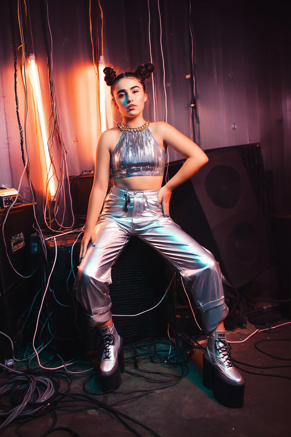 Woman in a metallic 90s pop star Halloween costume with platform shoes and baby buns, posing backstage with neon lights