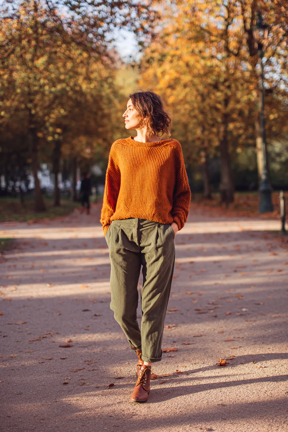 Woman walking through autumn park in burnt orange sweater and olive green pants, showcasing earthy tones in fall fashion trends