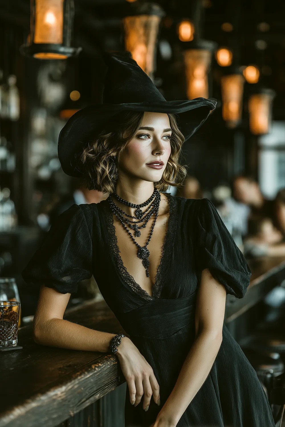 Woman in a modern witch Halloween costume with black dress and wide-brim hat, leaning on a bar counter with moody lighting