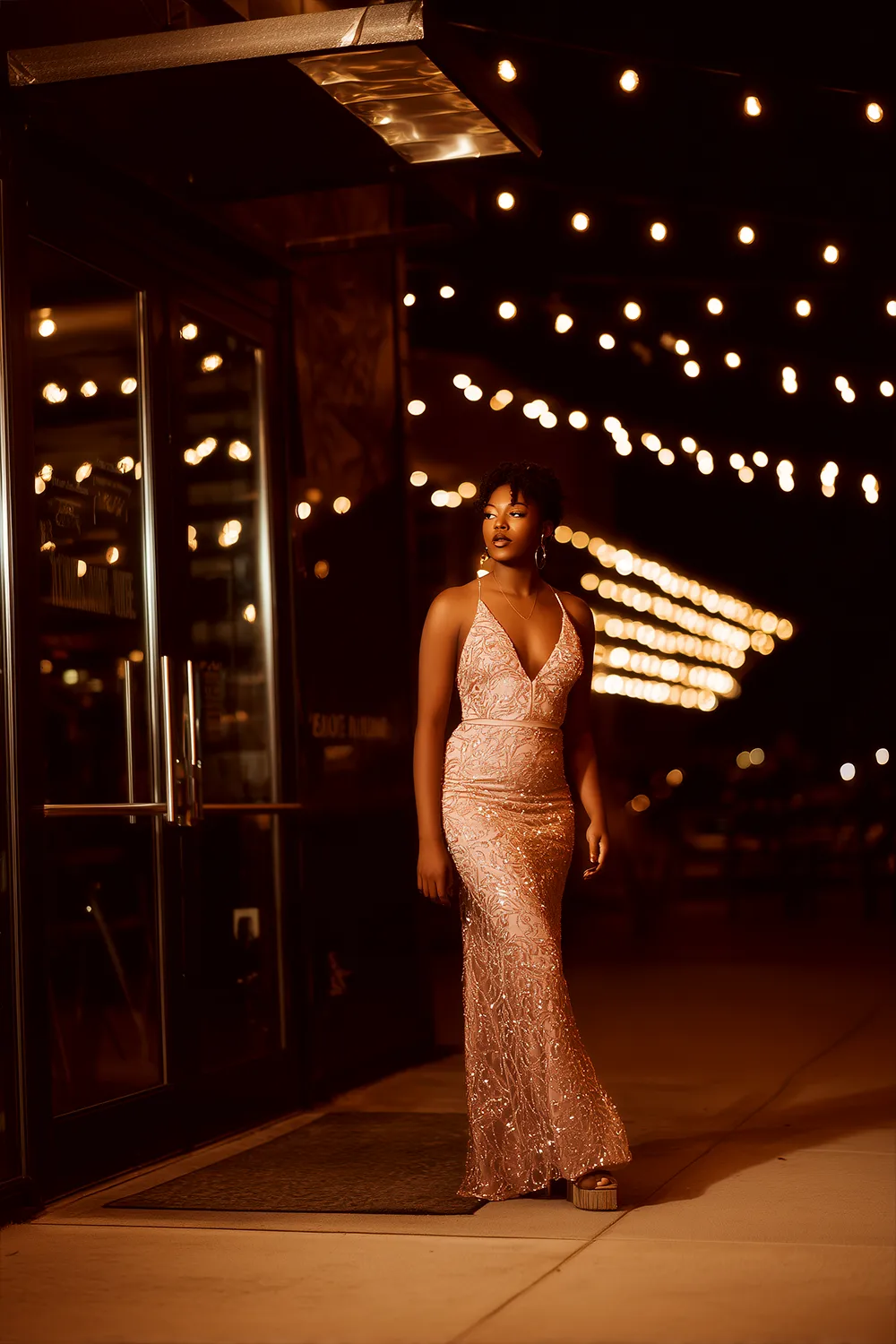 Petite woman in elegant V neck prom dress walking outside venue with string lights at night