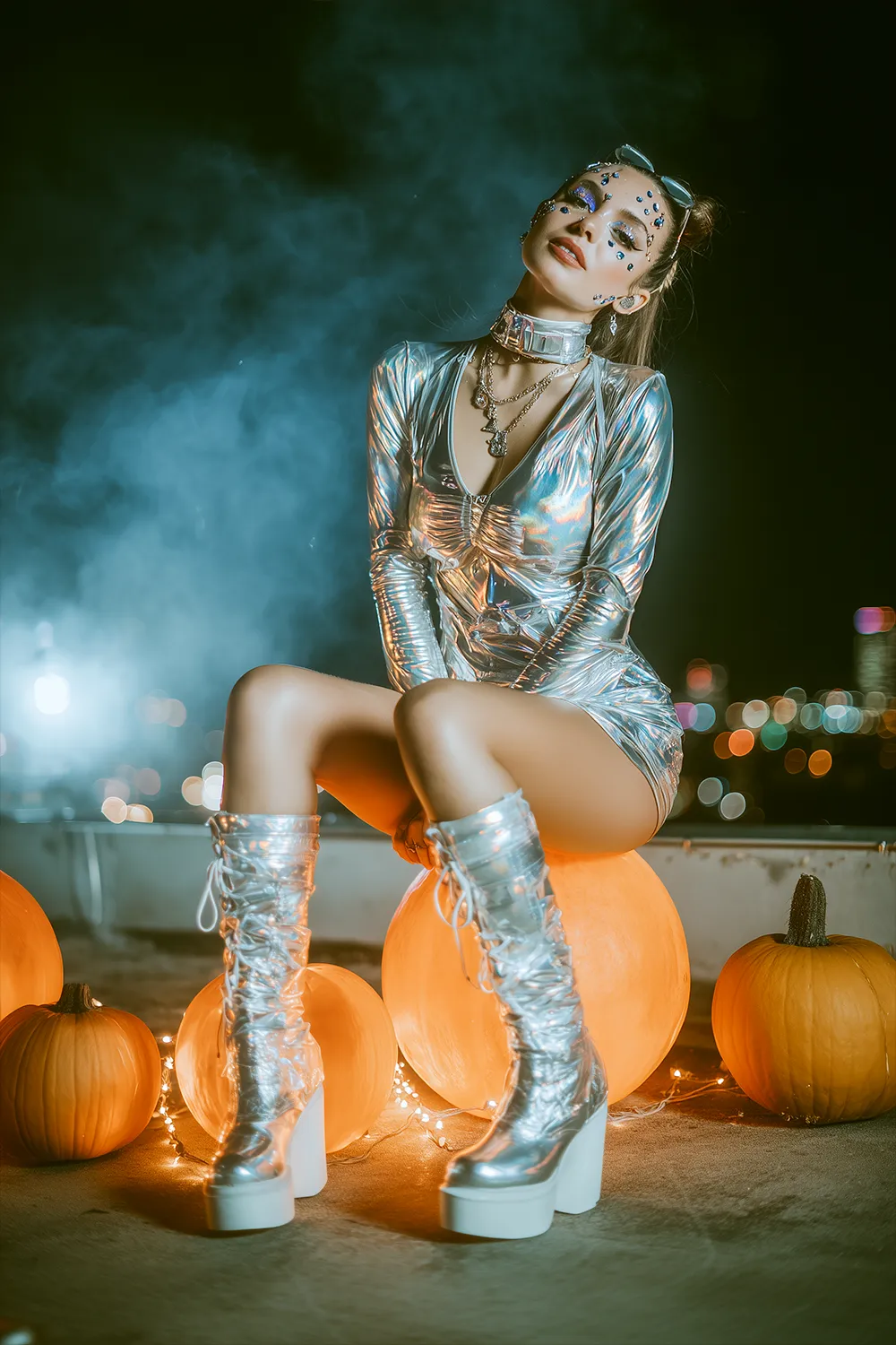 Woman in a metallic space babe Halloween costume with rhinestones and platform boots, sitting on pumpkins with city lights and fog