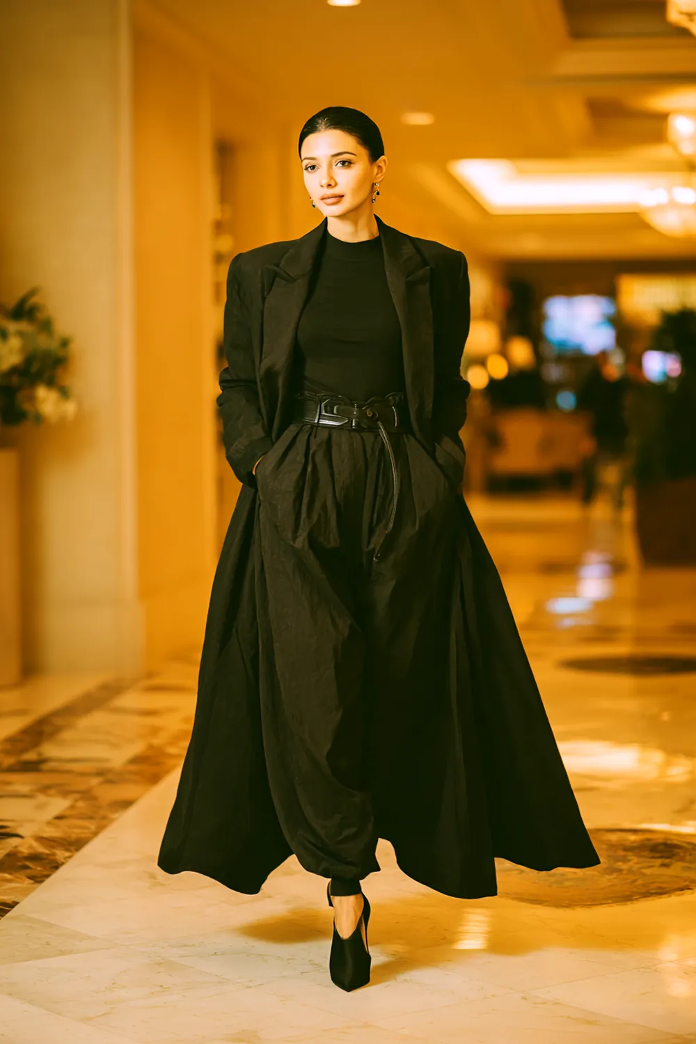 Woman in black cocktail dress styled with long coat walking through elegant hotel hallway